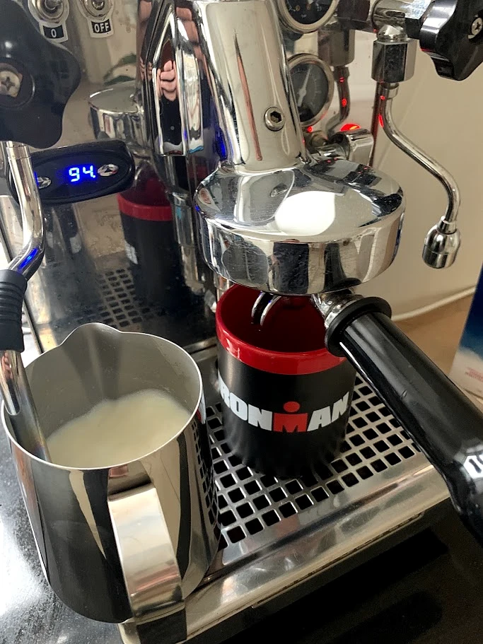 A milk pitcher beside a chrome espresso group, a thermometer reading 94 and a red-and-black mug